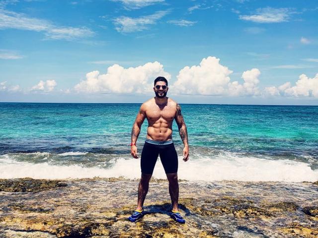 Muscular Lukas stands on rocky shore, wearing black shorts and blue flippers, with a blue ocean and white clouds in the background, showing his defined abs and tattoos.