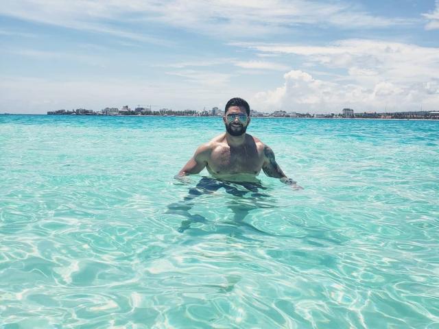 Lukas, shirtless, with a beard and tattoos, wading in clear blue water, smiling, wearing dark sunglasses, showing a muscular chest and arms, against a bright sky.