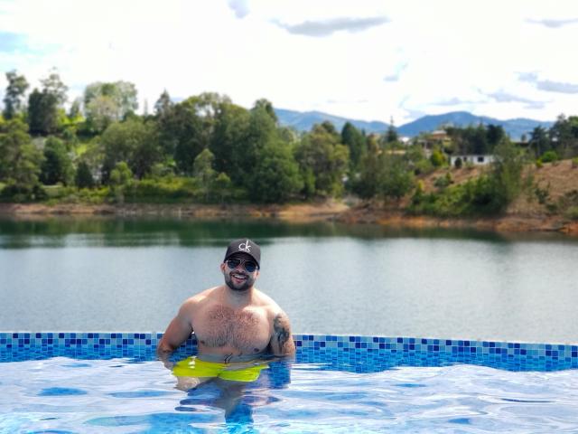 Lukas stands in a blue-tiled swimming pool, chest-deep in water. He's shirtless, with a hairy chest and wearing a black cap with a white logo. He has a beard and is smiling. A yellow inflatable ring is near his waist. The water reflects the bright sky and surrounding greenery. Lukas has tattoos on his right arm. The background shows a clear, blue sky with trees and hills. The image is sharp with natural light.