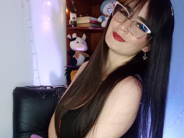 EllieBonneth with long brown hair, glasses, red lipstick, black tank top, and star earrings, smiling at the camera, showing her tits, in a cozy room with a stuffed toy.