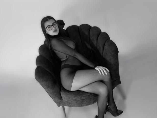 IrisBlair with glasses, dark lipstick, and long black hair, sitting on a dark, velvet chair, wearing a sheer, long-sleeve, bodysuit, exposing her tits and ass.