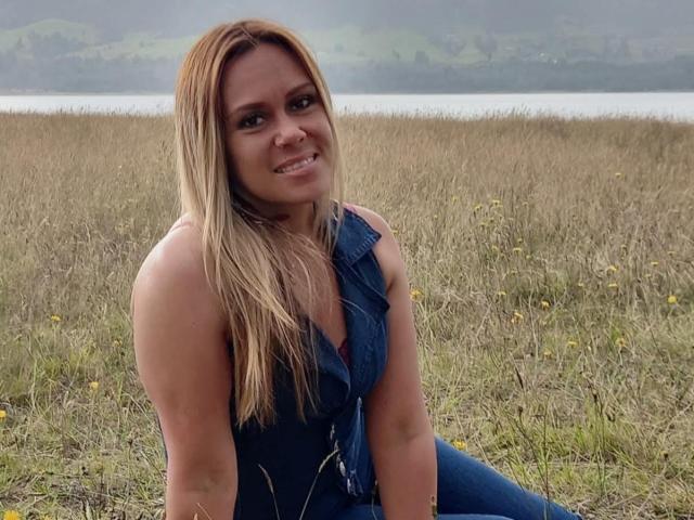LilithBonneth sits in a grassy field, looking at the camera with a slight smile. She has long, straight blonde hair and wears a sleeveless blue denim vest over a red shirt. Her skin is fair, and she has a slightly athletic build. The grass is brown and dotted with small yellow flowers. The background is misty with a lake and green hills. The composition is centered on LilithBonneth, who is slightly leaning forward. The image has a natural, candid feel.