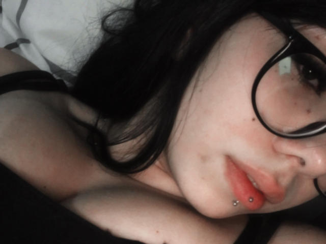 RavenahXz has pale skin, black hair, glasses, a nose piercing, and a lip piercing. Her tits are partially visible, and she's lying down with a black top.