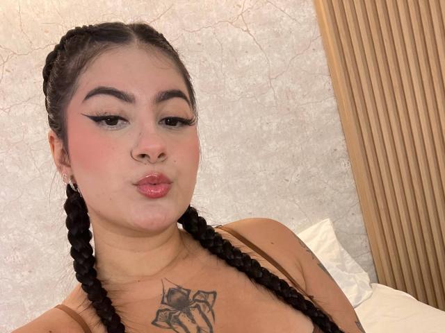 AlanaaWalker has black braided hair, full pink lips, and a tattoo of a bow on her chest. She's wearing a brown strap, looking at the camera with a pout.
