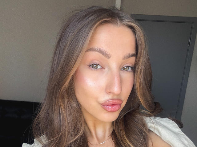 Kitkata has shiny brown hair, green eyes, and glossy pink lips. She's looking at the camera with a slight smile. Her white shirt is visible.