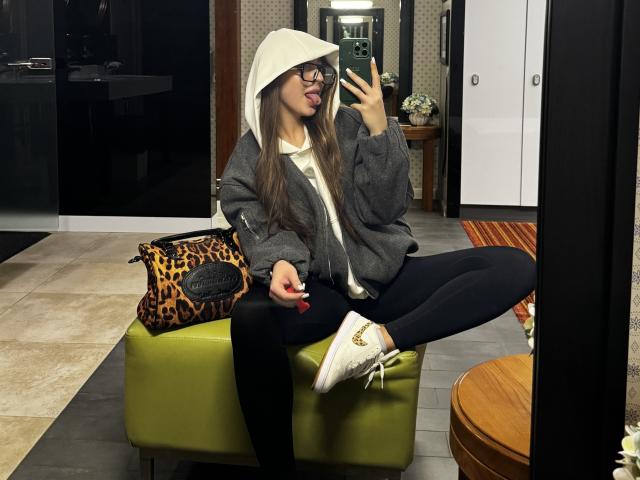 Kitkata, tongue out, wearing glasses, white hoodie, gray jacket, black pants, white sneakers, sitting on green chair, leopard bag, phone in hand, long brown hair, mirror selfie.