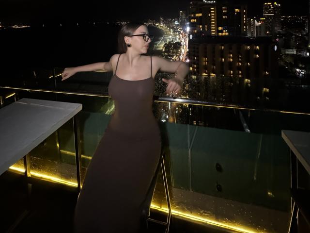 Night photo of Kitkata with glasses, brown dress, large tits, leaning on glass railing, city lights in background, black hair, yellow lighting, standing with arms out, dark setting.