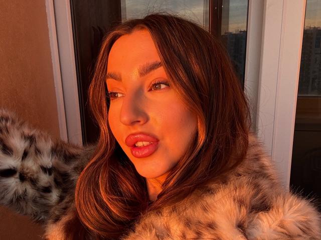Kitkata with long brown hair, orange sunlit skin, and pink lips, wearing a furry coat, has her arm raised, looking to the side with a slight pout.