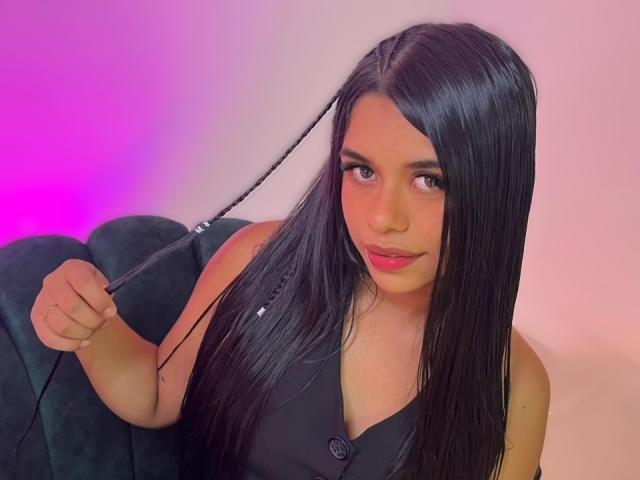 NoemieBlanc with long black hair, holding a black braid in her right hand, wearing a black sleeveless top, pink and purple lighting, sitting on a green velvet couch.