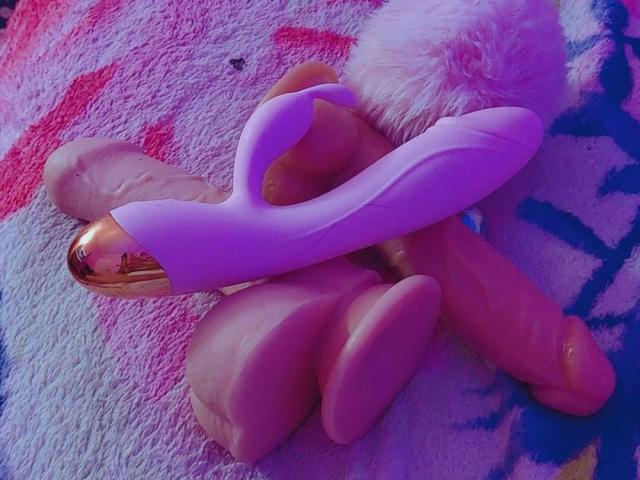 AngellVisen's pink dildo with a shiny gold tip is on top of a pair of realistic, flesh-colored testicles, resting on a fuzzy white and pink blanket.