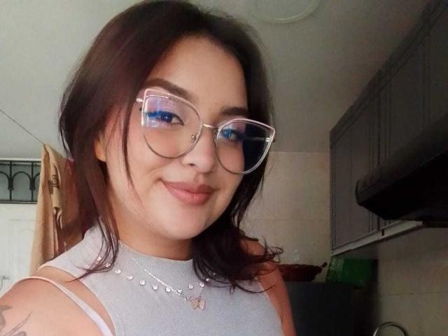 MissSubmissive mit mittelbraunen Haaren, die eine Brille trägt, ein graues Top ohne Ärmel und eine Halskette, lächelt in die Kamera und zeigt ein Tattoo auf ihrem rechten Arm..