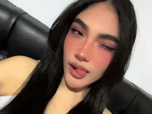 Miss Submissive hat langes, schwarzes Haar, rosigen Lidschatten und gerötete Wangen. Sie hat ihre Augen leicht geschlossen und die rosa Lippen sind leicht geöffnet, während sie auf einem schwarzen Ledersofa sitzt..