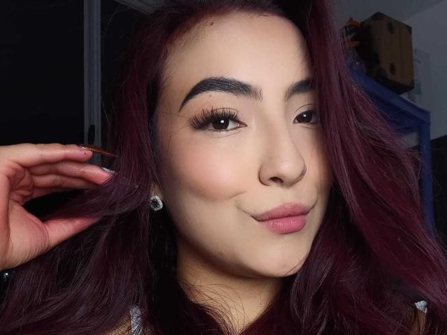 SilvanaGoddess has fair skin, dark red wavy hair, thick eyebrows, long eyelashes, pink lips, and is smiling with a hand in her hair, showing off her diamond earrings.