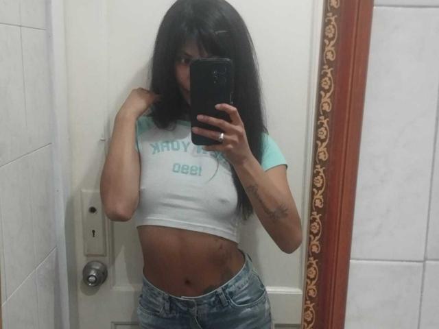 StormyWest with long black hair, wearing a white-green crop top and blue jeans, showing a tattoo on her right arm, taking a mirror selfie with her phone.