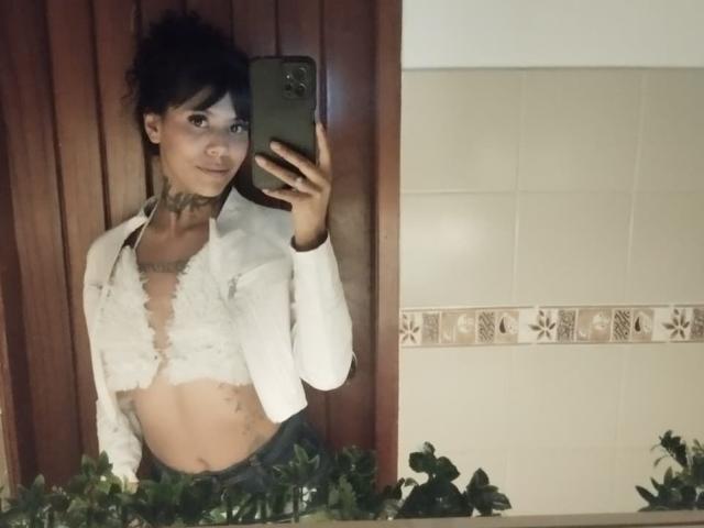 StormyWest with messy dark hair, wearing a white fluffy jacket, exposing her small tits, holding a phone, in a bathroom mirror selfie. She's wearing a choker and green pants.