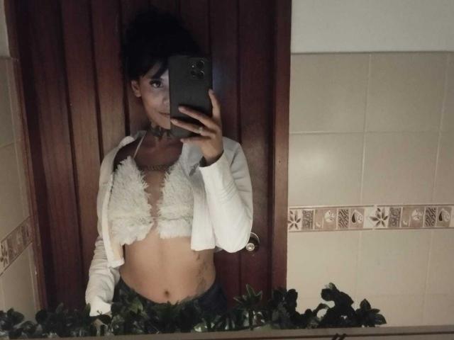 StormyWest taking a mirror selfie, wearing a white fluffy bralette, open white cardigan, black choker, and green leaf garland, showing a flat stomach and small tattoos.