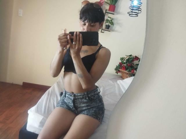 StormyWest with short black hair, wearing a black bra and denim shorts, taking a mirror selfie. She has tattoos on her arms and is holding a black phone.