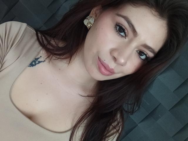 LindaMancini has light skin, brown hair, blue eyes, and a small lip piercing. She's wearing a beige top, showing a small tattoo on her right chest.