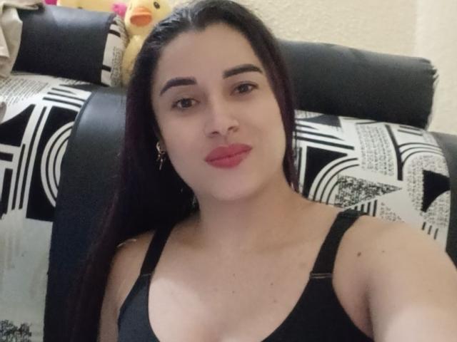 HanaMariana with dark hair, wearing a black tank top, showing her boobs, red lipstick, and earrings, sitting on a black and white patterned couch.
