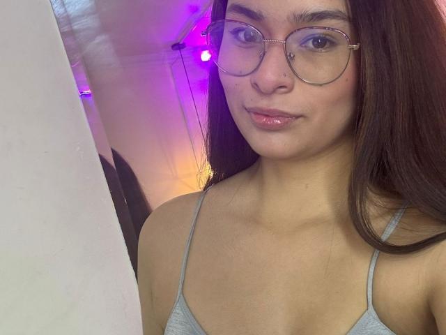 IsaDoux with glasses, brown hair, and a grey tank top, showing her tits and a small nose piercing, smiling slightly. Background has purple and yellow lights.