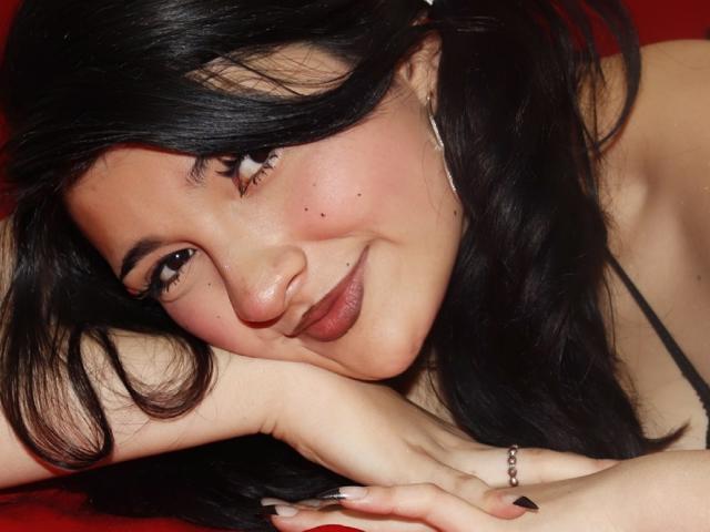 SweetEescape has black hair, brown eyes, and a cute smile with a slight blush. She's lying down, resting her head on her arm, showing her tits and a ring.