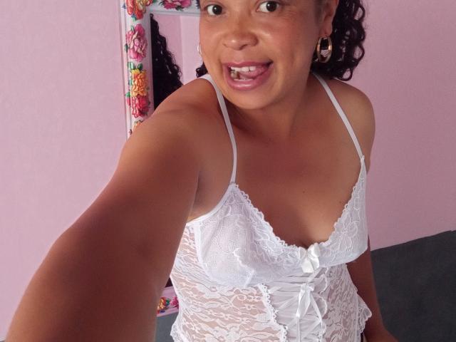 Rahel69 with curly brown hair, wearing a white lace bra, smiling with teeth showing, holding out arm for selfie, pink wall in background, floral frame on left.