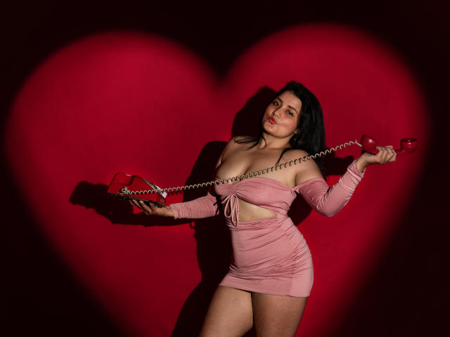 VelvetRosse with black hair, pink off-shoulder mini-dress, holding red phone with coiled cord, standing, shadowed by large red heart, looking sultry, showing her ass, in dim light.