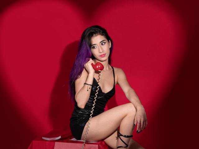 JuliaJobs with purple hair, black shiny dress, holding red phone, sitting on red stool, legs crossed, looking at camera, red heart shadow, coiled phone cord, sexy.