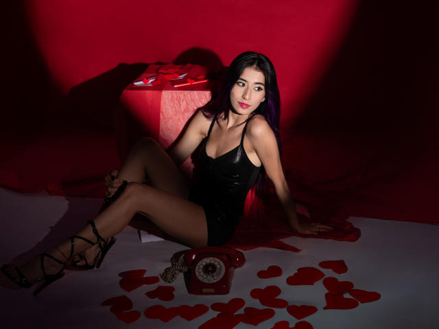 JuliaJobs sits against a red backdrop, wearing a black leather dress and strappy black heels. Her long dark hair has purple highlights. She leans back with her legs crossed, arms resting on the ground. A red telephone and scattered red heart shapes are on the floor. Her red lipstick matches the red lighting and backdrop, creating a sultry, intimate mood. The composition uses strong contrasts and shadows to highlight her figure and the objects. The overall style is provocative and sensual.