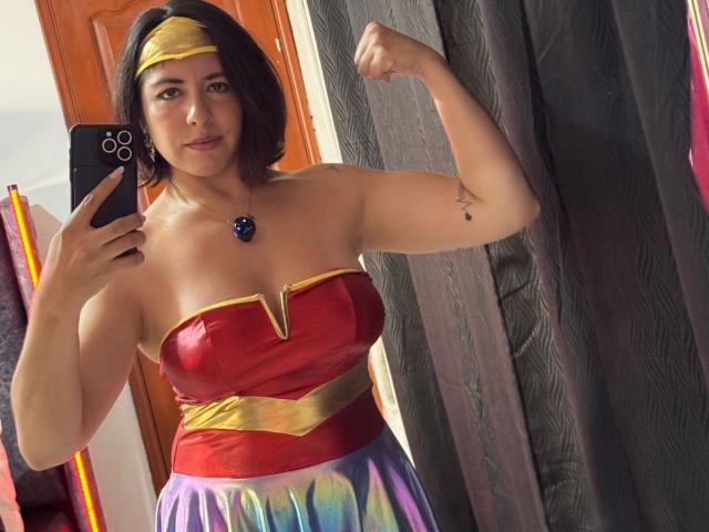 AnastasiaEvansX, curvy woman with medium tits, wearing Wonder Woman cosplay, red strapless top, yellow headband, flexing arm, holding phone, purple skirt, dark hair, tattoo on right arm.