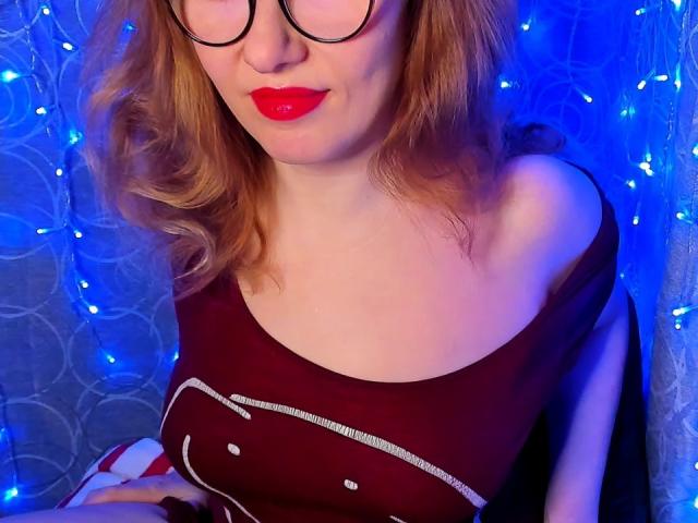 PaulinaSunny with red lipstick, glasses, and wavy blonde hair, wearing a red off-shoulder top, showing her bare shoulder and chest, surrounded by blue fairy lights.