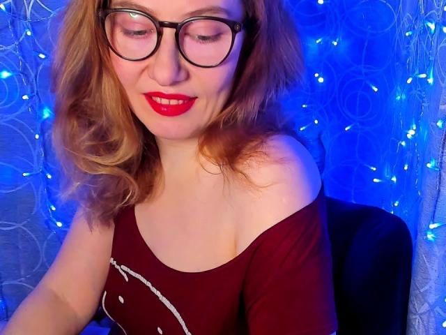 PaulinaSunny with wavy red hair wears black-rimmed glasses and bright red lipstick. She's in a maroon off-shoulder top with a white smiley face design. She looks down with a slight smile. The blue light creates a glow on her fair skin. String lights are in the background. Her hair is loosely parted. She's seated, and the image is focused on her upper body.