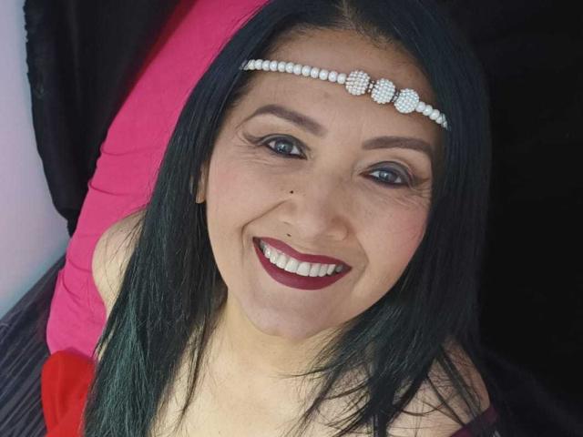 Close-up photo of SweetFoxyne with long black hair, smiling with red lipstick. She has a small black mole on her right cheek. She wears a white pearl headband with three round, diamond-like studs. Her eyebrows are neatly shaped, and she has dark eyeliner. She's looking up, slightly to the right. The background is mostly black with a pink pillow visible on the left. The lighting is even, highlighting her face.
