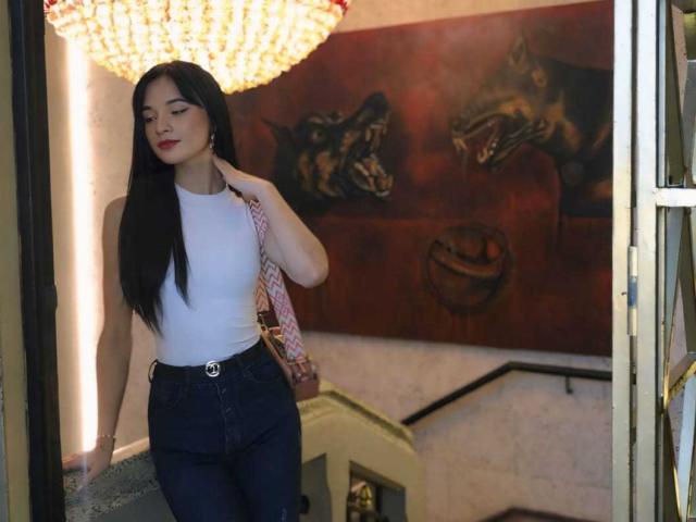 PamelaKander with long black hair, white sleeveless top, high-waisted blue jeans, holding pink bag, standing near ornate chandelier, looking slightly to the side, smiling with red lips.