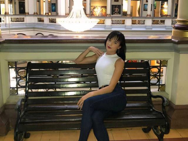 PamelaKander with black hair, white sleeveless top, and blue jeans sits on a black metal bench, hands near neck, looking at camera, in a well-lit indoor setting.