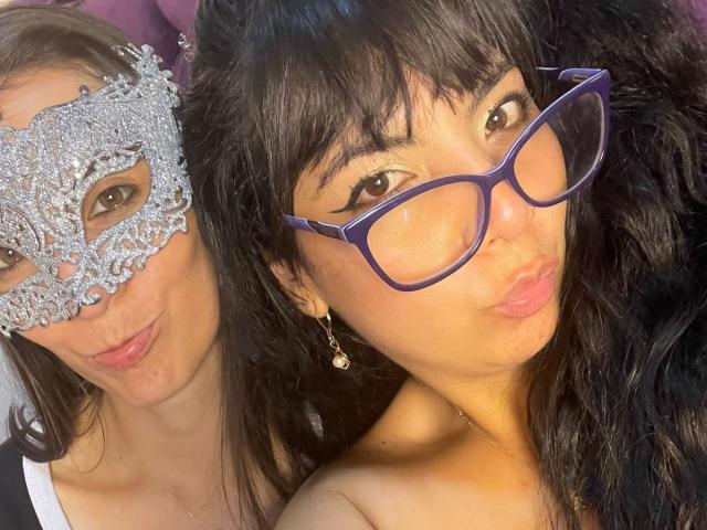 Two women in a close-up selfie. ValkiriaAndDione on the left wears a silver, ornate masquerade mask with intricate lace patterns, her brown hair is straight and loose. ValkiriaAndDione on the right has dark, wavy hair and wears purple glasses, making a pouty face. Both have light skin and are closely positioned. ValkiriaAndDione on the right has gold hoop earrings and a slightly tanned complexion. The image is brightly lit with a focus on their faces.