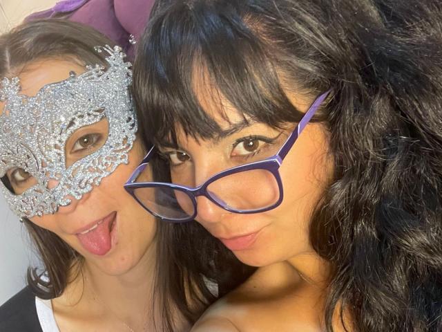 Close-up of two women. ValkiriaAndDione on the left wears a silver lace masquerade mask with intricate patterns, sticking out her tongue. She has light brown hair and fair skin. ValkiriaAndDione on the right has dark, curly hair, wears purple-framed glasses, and looks at the camera with a slight smile. Both have brown eyes. The image focuses on their faces and masks, with ValkiriaAndDione on the right slightly closer to the camera. The right woman's glasses contrast with her dark hair.