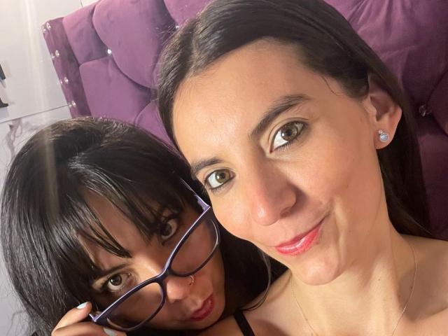 ValkiriaAndDione with glasses, cute brown eyes, and dark hair, sitting next to ValkiriaAndDione with long dark hair, pink lipstick, and diamond earrings, smiling at the camera.