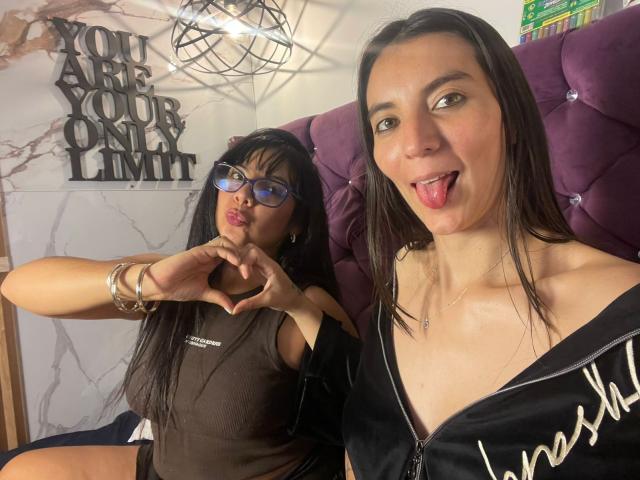 ValkiriaAndDione with glasses, making a heart shape with hands, brown top, ValkiriaAndDione with tongue out, black zip-up, purple headboard, and metal wall decor. Both have long black hair.