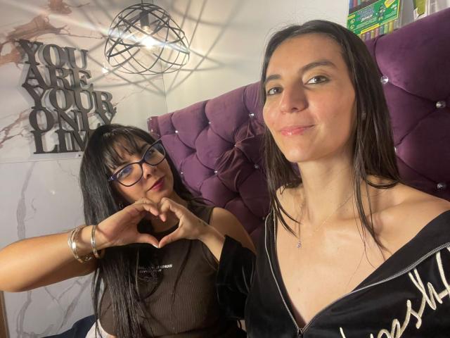 ValkiriaAndDione with glasses, making a heart shape with her hands, next to ValkiriaAndDione with a smile, wearing a black dress, in a bedroom with purple headboard.