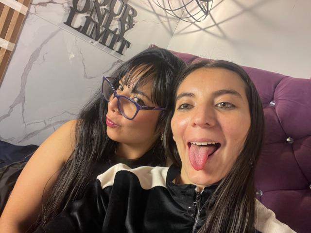 ValkiriaAndDione with black hair and glasses, pouting, has small tits and dark eyes. Dione with brown hair, tongue out, has medium tits and dark eyes, wearing a black and white shirt.