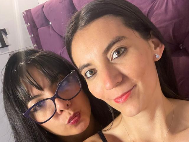 ValkiriaAndDione with glasses, puffy tits, and dark hair, and ValkiriaAndDione with brown eyes, red lips, and straight hair, on a purple tufted couch.