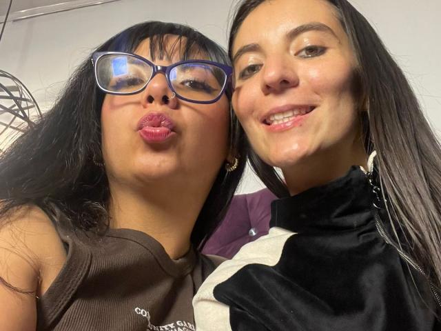 Two women in a close-up selfie. ValkiriaAndDione on the left has dark brown skin, black hair, and wears blue glasses. She pouts her lips with pink lipstick. ValkiriaAndDione on the right has fair skin, black hair, and smiles with visible braces. She wears a black and white jacket. The image is bright with clear, sharp details. The right ValkiriaAndDione's jacket contrasts with her skin, while the left ValkiriaAndDione's pink lips stand out. The composition focuses on their faces.