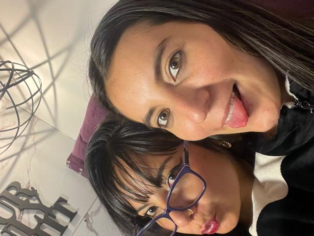 Close-up selfie of ValkiriaAndDione with two faces. ValkiriaAndDione has straight black hair, brown eyes, and is sticking her tongue out. The second ValkiriaAndDione has black-framed glasses, straight black hair with bangs, and is pouting. Both have fair skin. ValkiriaAndDione on the left wears a black jacket, the one on the right a white shirt. The lighting is even, highlighting their facial features. The image is sharp with a playful, mischievous vibe.