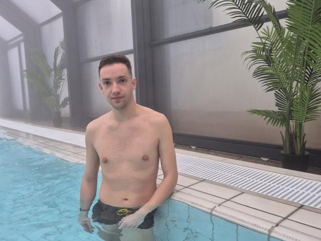 Cocco69 is standing in a pool, shirtless, with a slight mustache, and wearing black swim trunks, showing a flaccid, uncircumcised penis. His skin is fair, and he's smiling.
