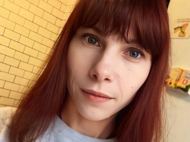 KateFire with red hair, blue eyes, and light skin, wearing a white shirt, has a slight smile, showing subtle freckles and a small beauty mark under her right eye.