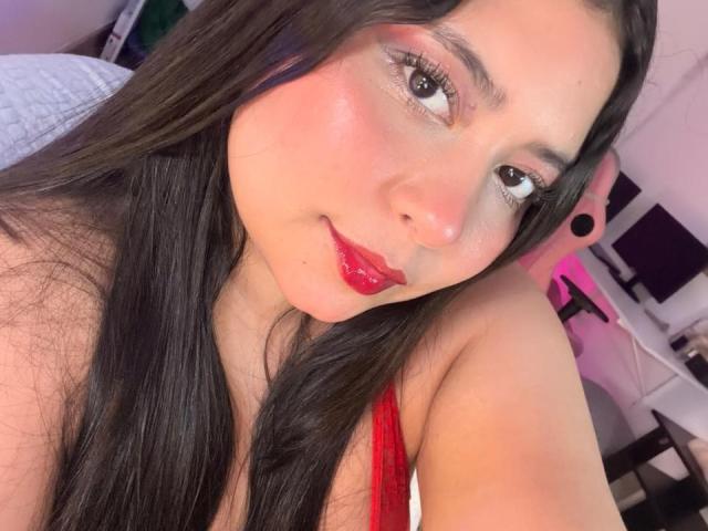 DakotaJhons69 with long black hair, red lipstick, and pink eyeshadow, smiling at the camera, showing her red top and bare shoulders, with a cute and sexy look.