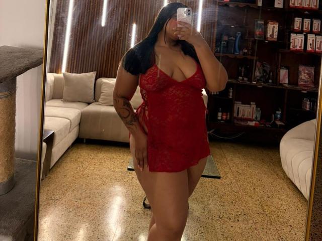 DakotaJhons69 in red lace lingerie, showing her tits and ass, with long black hair, tattoo on arm, taking a mirror selfie, holding a phone, in a living room.
