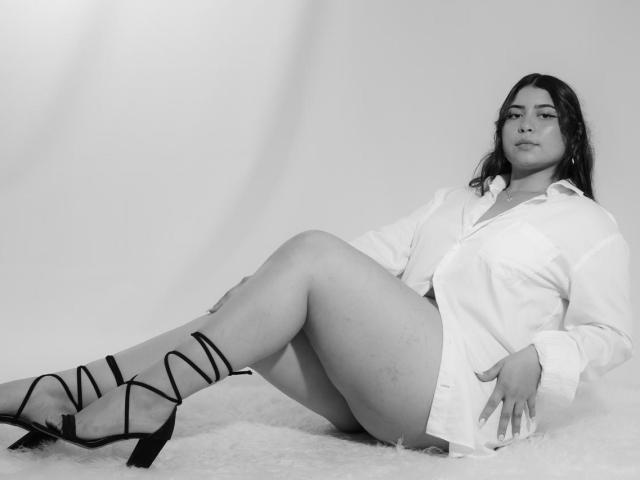 DakotaJhons69 sitting on a fluffy rug, legs crossed, wearing a white shirt, black strappy heels, looking confident, slightly chubby with long dark hair, no pants.