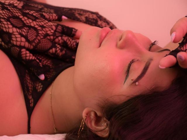 DakotaJhons69 with purple hair, black lace top, and pink nail polish, lying down, eyes closed, hand on forehead, gold hoop earrings, nose piercing, pink lighting, relaxed expression.